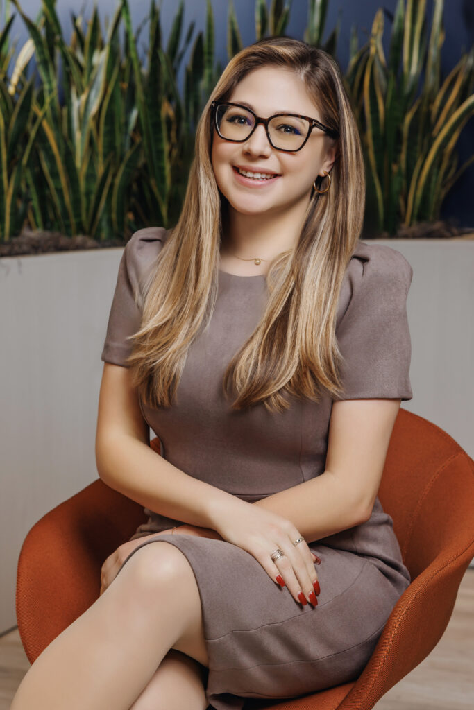 Salina Kelley sitting on a dark red chair with plants on the background in an office setting, dressed professionally and smiling to camera.